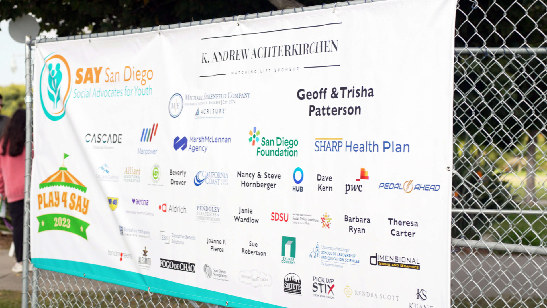 Banner titled "SAY San Diego Social Advocates for Youth" displayed on a chain-link fence, showcasing various sponsors through a corporate partnership in San Diego, including companies and individuals.