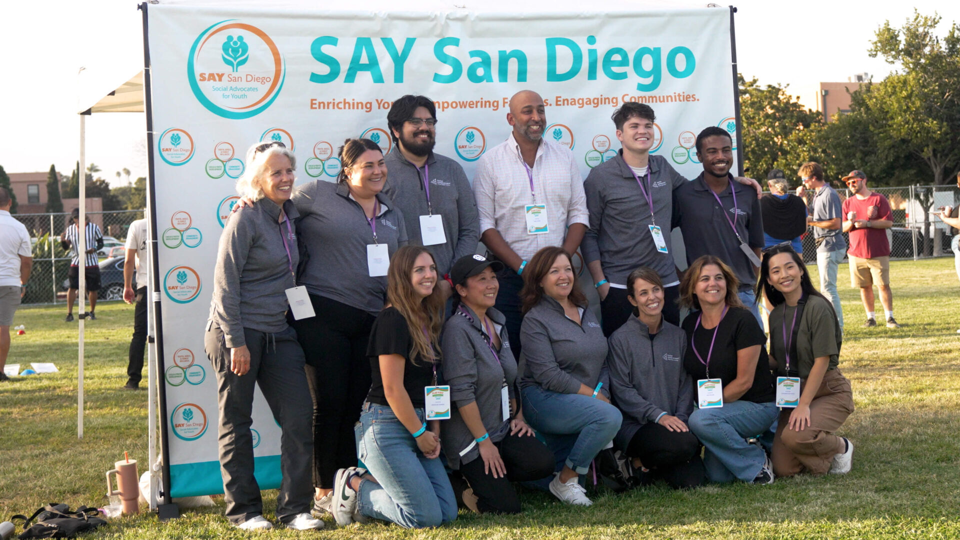 A group of people poses in front of a "SAY San Diego" banner on a grassy field, showcasing their Corporate Partnership in San Diego. They are wearing matching shirts and name tags, smiling for the photo. Trees and other people are visible in the background.