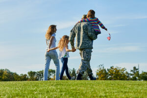 A soldier in uniform walks on a grassy field holding hands with two children. Another child wearing colorful clothing rides on the soldier's shoulders, holding a small flag. An adult walks alongside them.