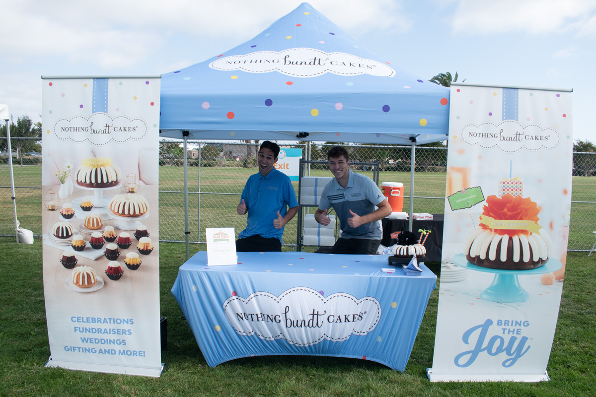 Two people stand under a blue "Nothing Bundt Cakes" tent at an outdoor Play 4 SAY event. The table displays various cakes with promotional banners on either side.