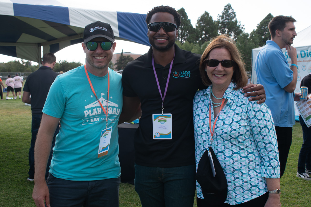 Three people smiling and posing together outdoors, two men and one woman, wearing casual clothes and event badges around their necks. They are standing on a grassy area with a tent in the background, clearly enjoying themselves at the Play 4 SAY event.