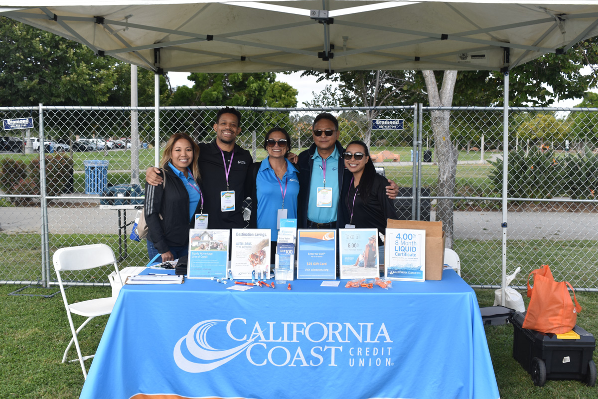 Five people standing behind a table with a California Coast Credit Union tablecloth at an outdoor event. The table displays informational brochures, promotional items, and details about the Play 4 SAY initiative.