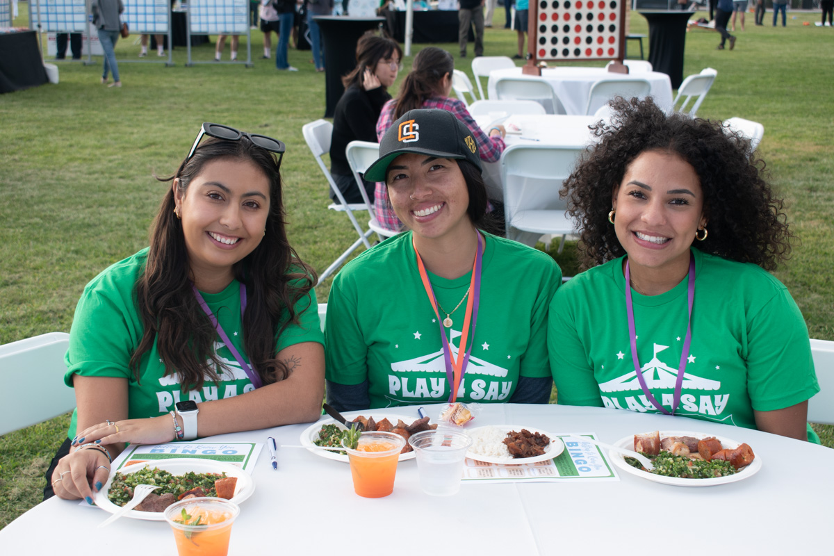 Three people wearing green "Play 4 SAY" shirts sit at a table with plates of food and drinks during an outdoor event. There are other attendees and event booths visible in the background.