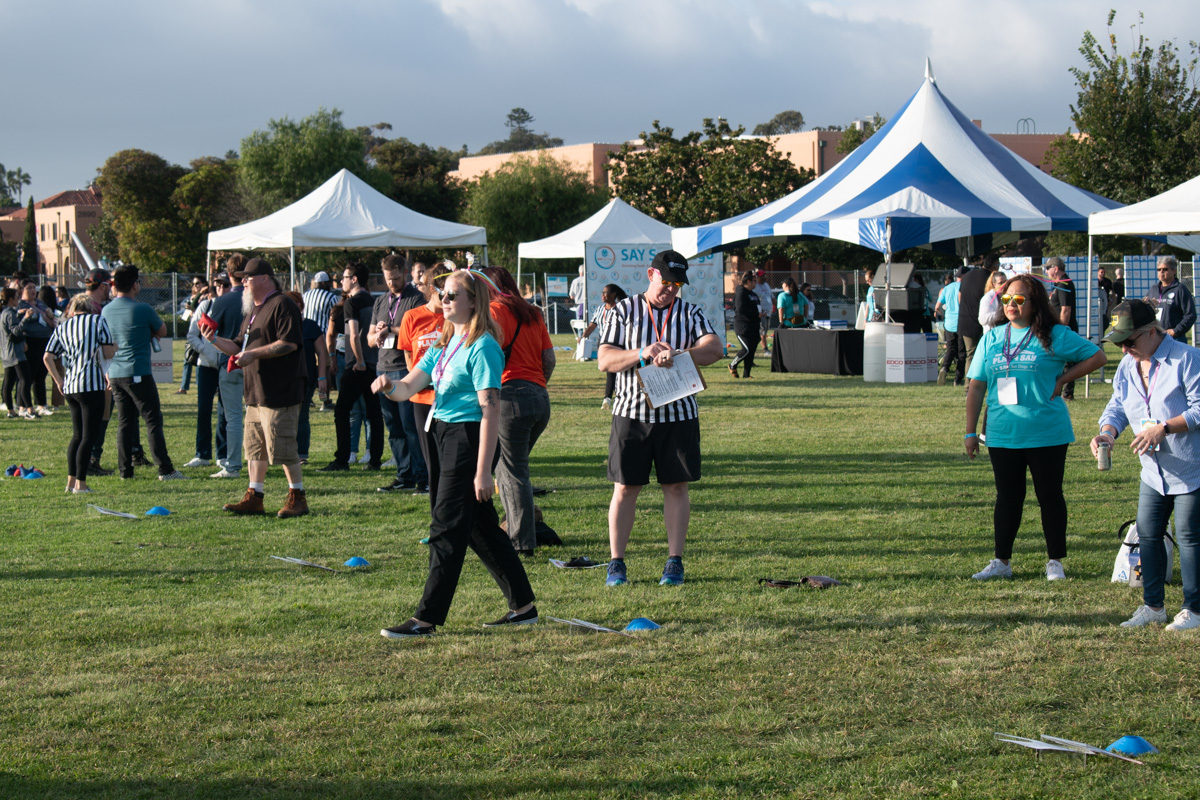 A group of people participates in the Play 4 SAY event on a grassy field, with several white and blue tents in the background. Some individuals are wearing referee shirts.