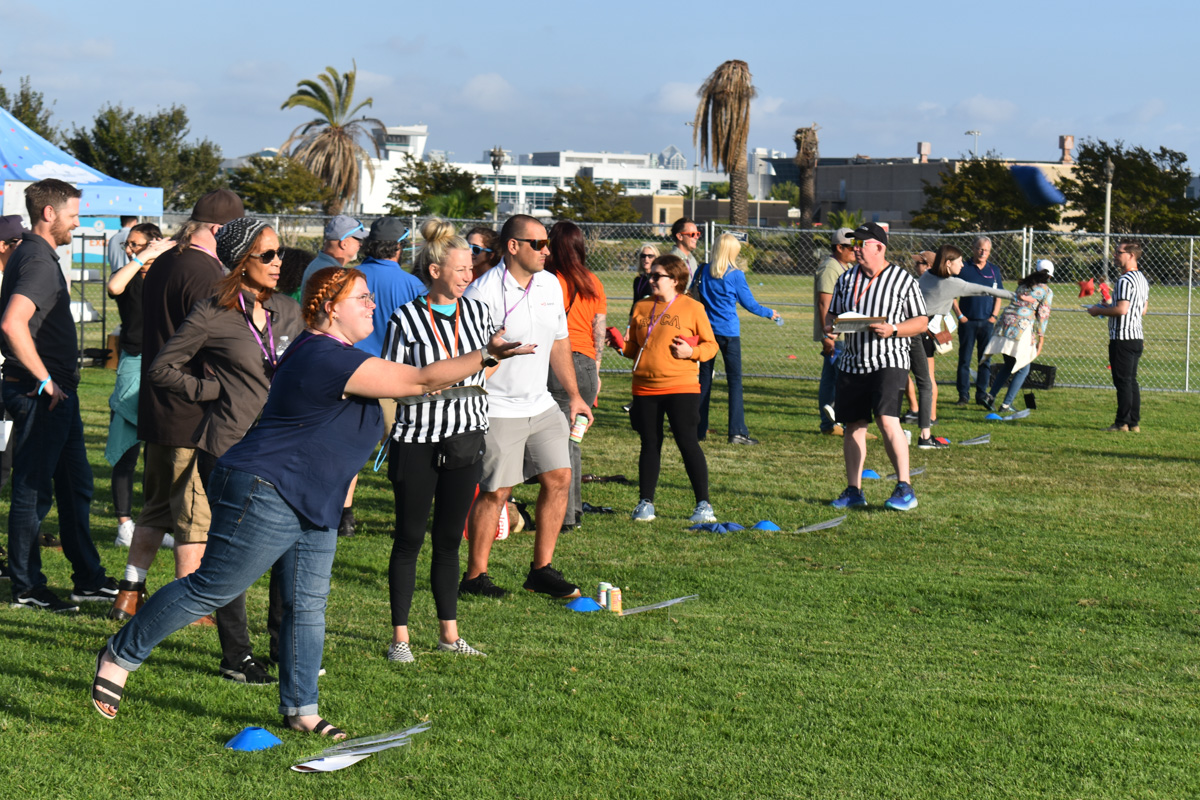 Outdoor field event with people participating and spectating. A woman in a striped shirt is throwing an object as part of the Play 4 SAY initiative while others, some in similar shirts, watch. Trees and buildings are visible in the background.