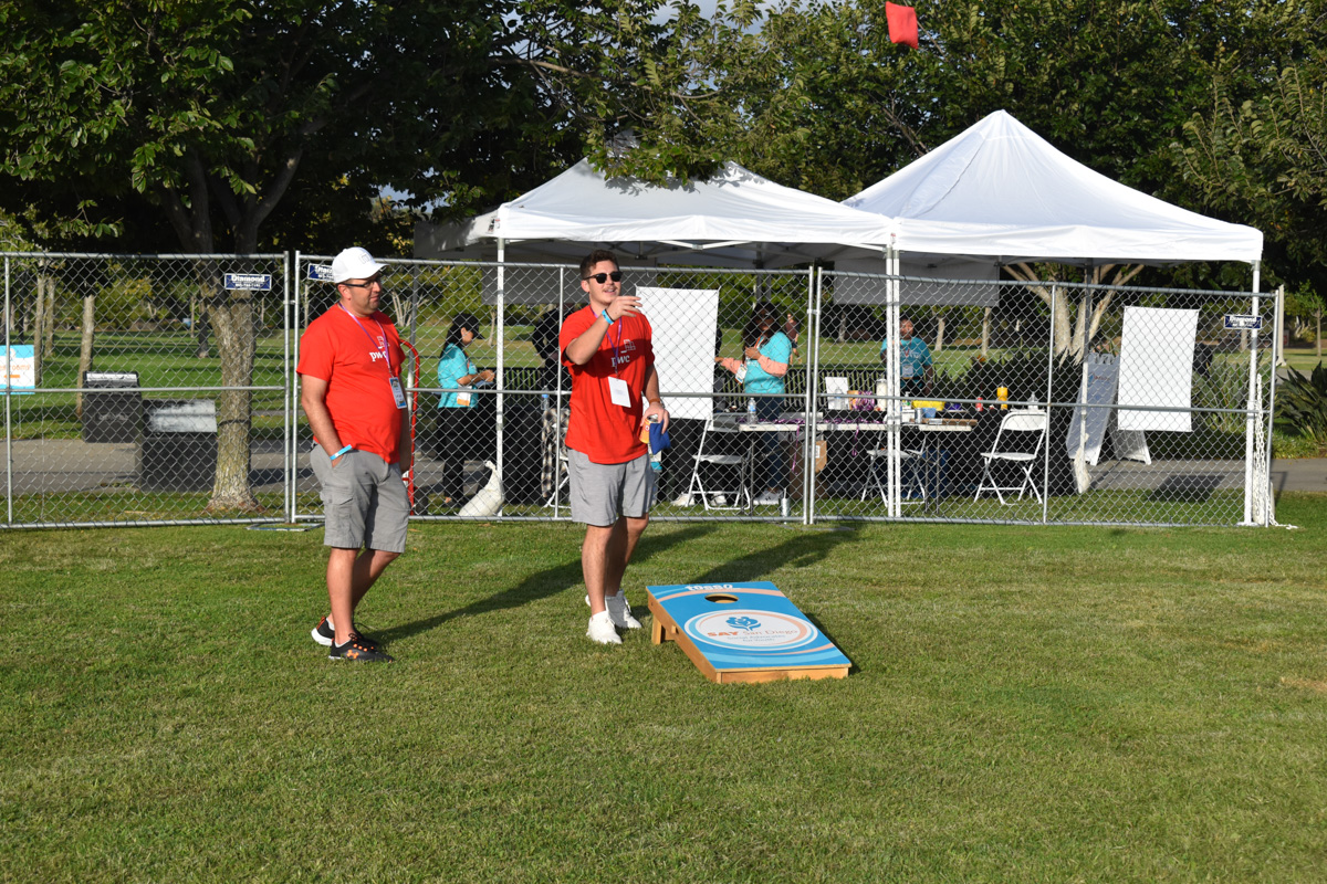 Two people in red shirts are playing cornhole on a grassy area in front of a fenced white tent setup at an outdoor event for Play 4 SAY.