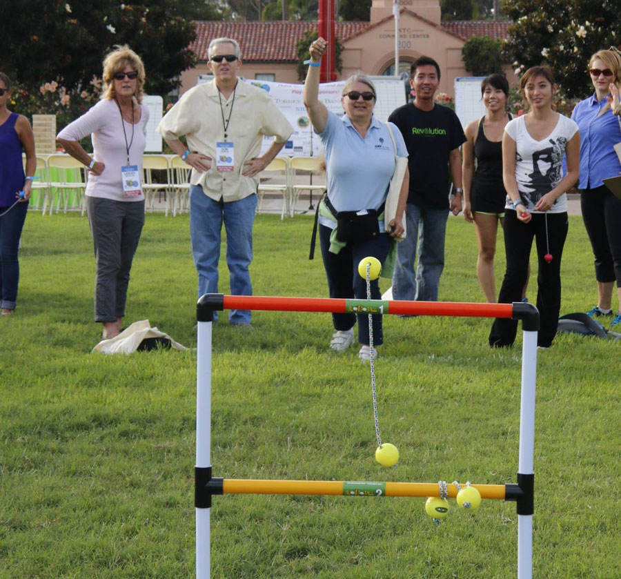 A group of people stand on a grassy field, with one person in the center throwing a bolos at a ladder toss game frame, while others discuss ways to donate the event's proceeds to charity.