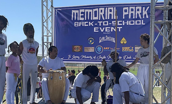 A group of individuals perform at the Memorial Park Back-to-School Event on an outdoor stage, accompanied by a large drum and a banner in the background displaying event and sponsor information.