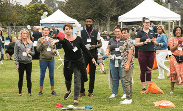 A group of people stand and watch as a woman throws a beanbag in an outdoor setting with tents and chairs in the background, embodying the spirit of San Diego Events.