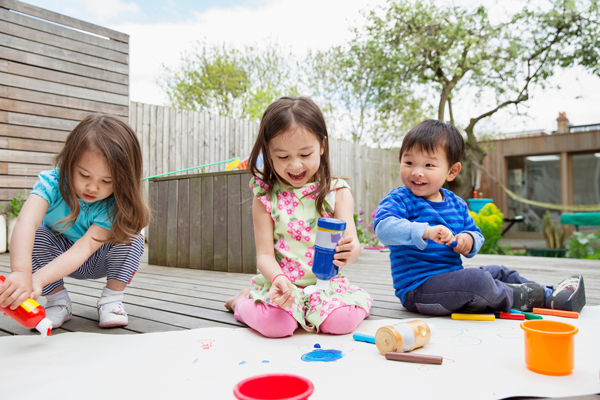 Three children are sitting and playing with colorful toys and markers on a wooden deck outside on a sunny day. They are smiling and appear engaged in their activities.