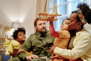 A family sits together indoors; an adult and two children are on the couch, with one child holding a plush toy and the other child holding a wooden airplane. Another adult kisses the child with the airplane.