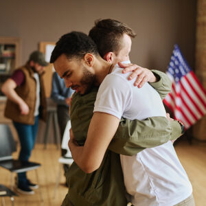 Two men are hugging each other in a well-lit room with wooden floors and American flag in the background. Another man in a brown jacket and green cap can be seen in the background.