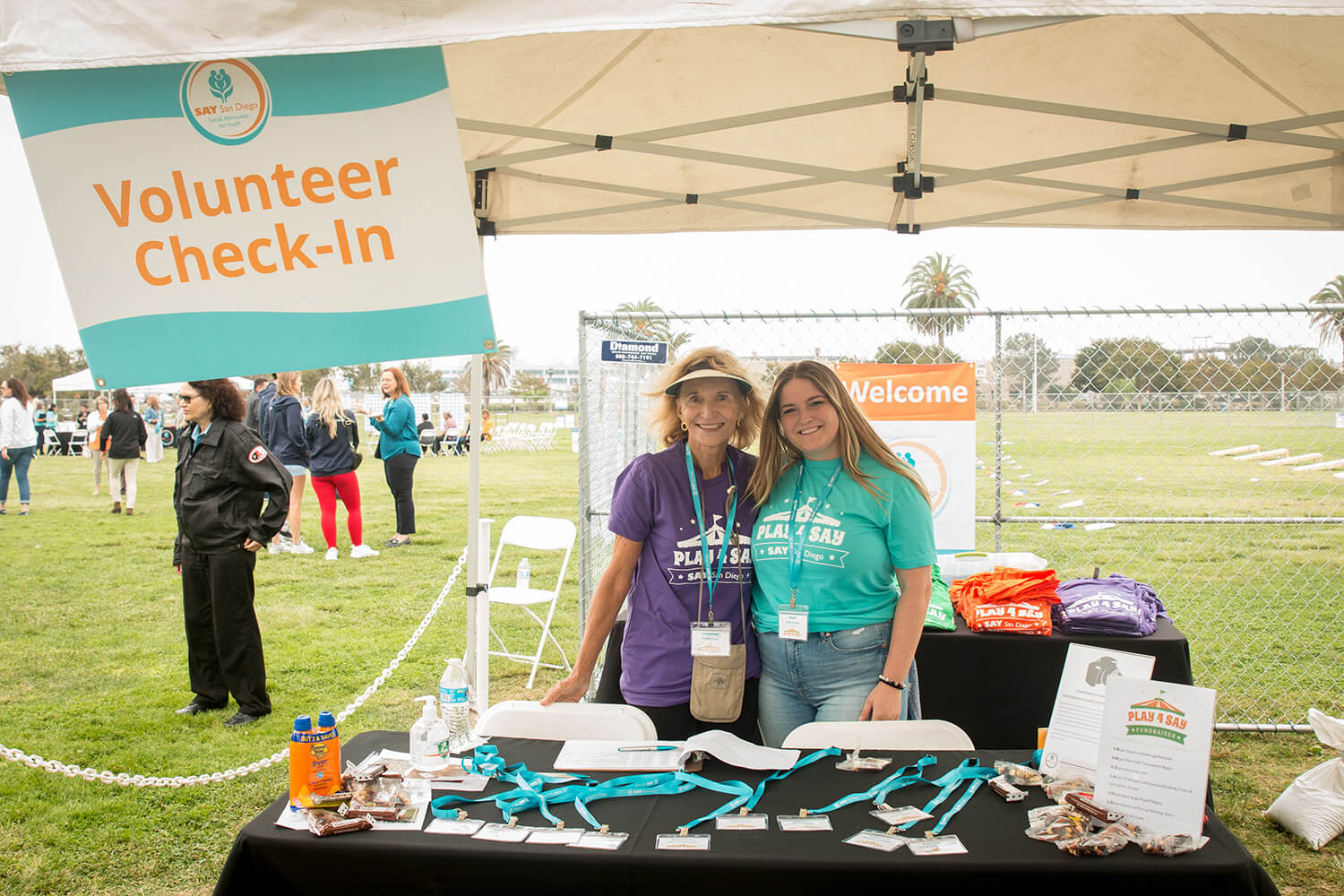 Two volunteers stand behind a check-in table under a tent labeled "Volunteer Check-In" at an outdoor San Diego event. The table holds name tags, snacks, and promotional materials. People and a grassy field are in the background.