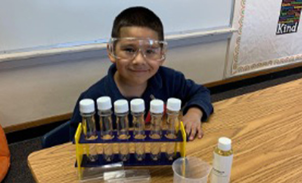 A young child wearing safety goggles sits at a classroom table with a rack of test tubes, a beaker, and a container of liquid in front of them, reminiscent of hands-on learning activities seen at San Diego events.