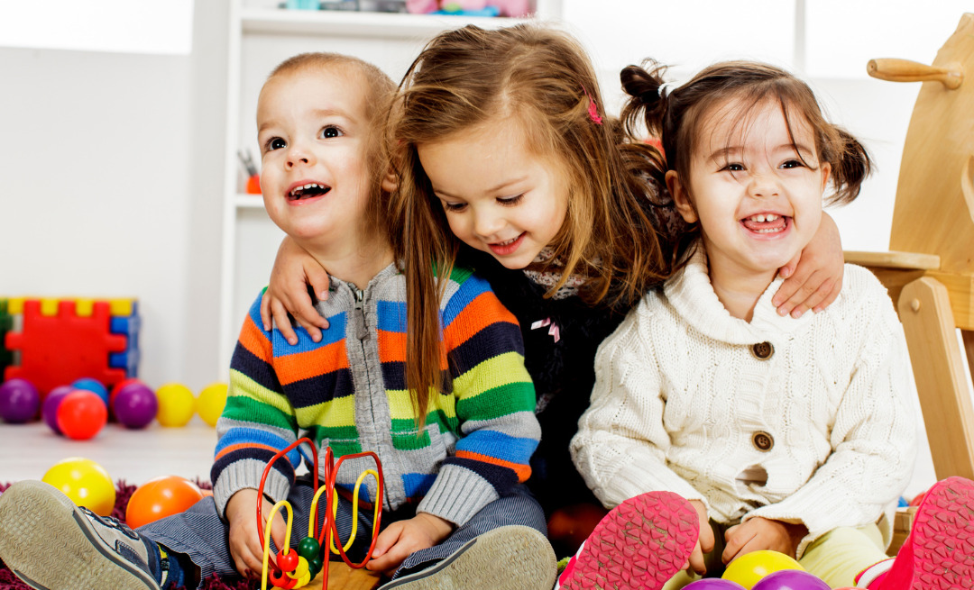 Three children are sitting on the floor, surrounded by colorful toys. One child is wearing a striped sweater, another is dressed in a dark top, and the third is in a white sweater. They are smiling and laughing.